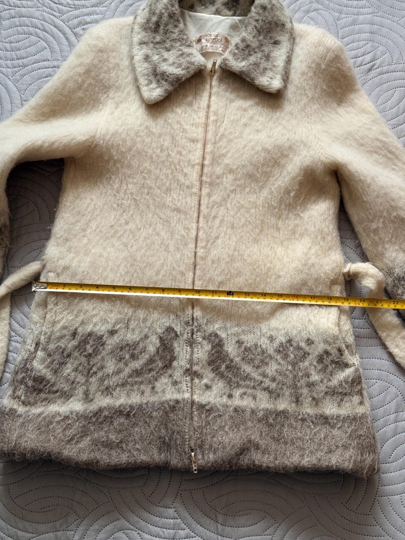 May include: Cream-colored zip-up coat with a faux fur collar and trim. The coat features a decorative pattern in shades of gray near the hem. Includes a matching belt. The coat measures approximately 30 inches in length.