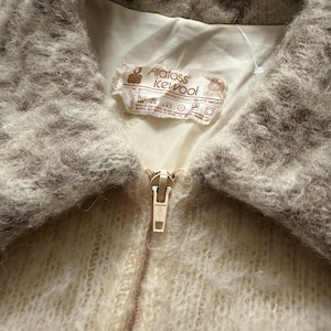May include: Close-up of a vintage cream-colored jacket with a fuzzy, textured collar and a gold zipper. The jacket has a label that reads "Alatoss Icewool". The collar is a mix of light and dark brown. The jacket is made of a soft, knitted material.
