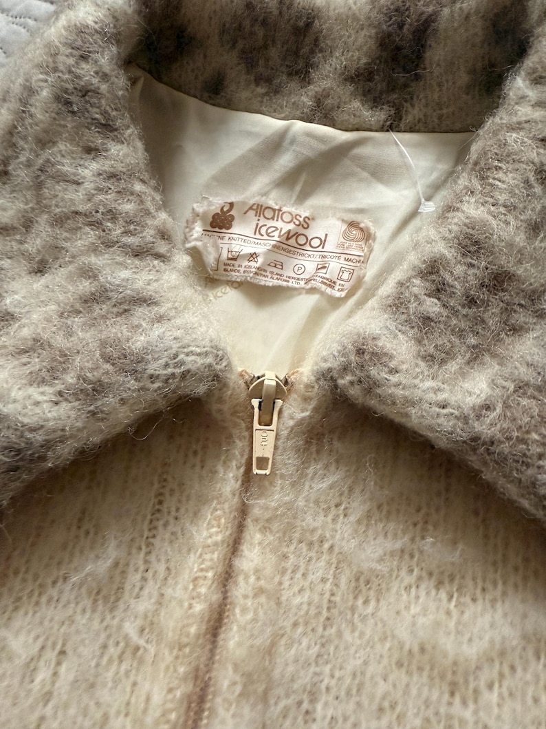 May include: Close-up of a vintage cream-colored jacket with a fuzzy, textured collar and a gold zipper. The jacket has a label that reads "Alatoss Icewool". The collar is a mix of light and dark brown. The jacket is made of a soft, knitted material.