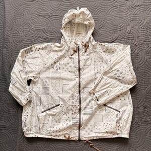 May include: White hooded jacket with a full-length zipper and a drawstring at the hood and waist. The jacket features a repeating pattern of gray geometric shapes and designs. The jacket has two front pockets and brown accents on the hood and zipper.