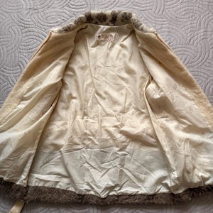 May include: Cream-colored vintage jacket with a brown fur trim around the collar and hem. The interior lining is a light cream color. The jacket has a matching belt and a label inside.