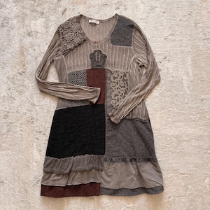 Op de afbeelding: Een patchwork jurk met lange mouwen, met een mix van texturen en kleuren. De jurk bevat verschillende stoffen, waaronder een kanten detail op de schouder en een geplooid zoom. Het kleurenpalet omvat tinten bruin, grijs, zwart en bordeaux.