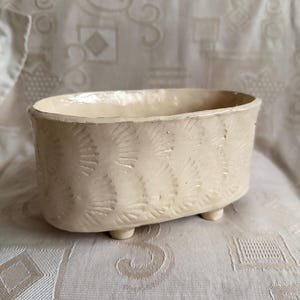 May include: Cream-colored ceramic planter with an oval shape and three small feet. The planter features a repeating fan-like pattern around the exterior. The interior is smooth, while the exterior has a textured design. A decorative item for plants.