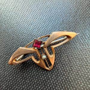 May include: A vintage gold-toned brooch with an Art Nouveau design. It features a central square red gemstone and a small pearl. The brooch has an asymmetrical shape with curved lines and pointed edges, and a pin closure.