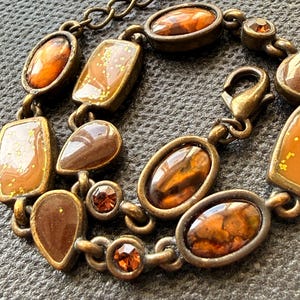 Könnte beinhalten: Ein Armband mit einer Reihe von bronzefarbenen Metallgliedern, die mit verschiedenen bernsteinfarbenen Cabochons in ovaler, rechteckiger und tropfenförmiger Form verbunden sind. Einige Segmente enthalten goldene Sprenkel, andere kleine runde Kristalle.