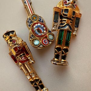 May include: Three decorative brooches: a mosaic guitar, and two gold-toned nutcracker soldier figures. The guitar brooch features a colorful mosaic design. The nutcracker brooches have red and blue accents, with rhinestone details.