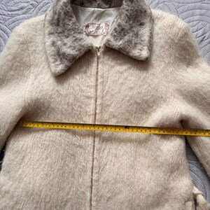 May include: Cream-colored vintage jacket with a fuzzy texture and a zip-up front. The collar and lower hem feature a patterned design in shades of gray. The jacket has a measurement of approximately 19 inches across the chest.