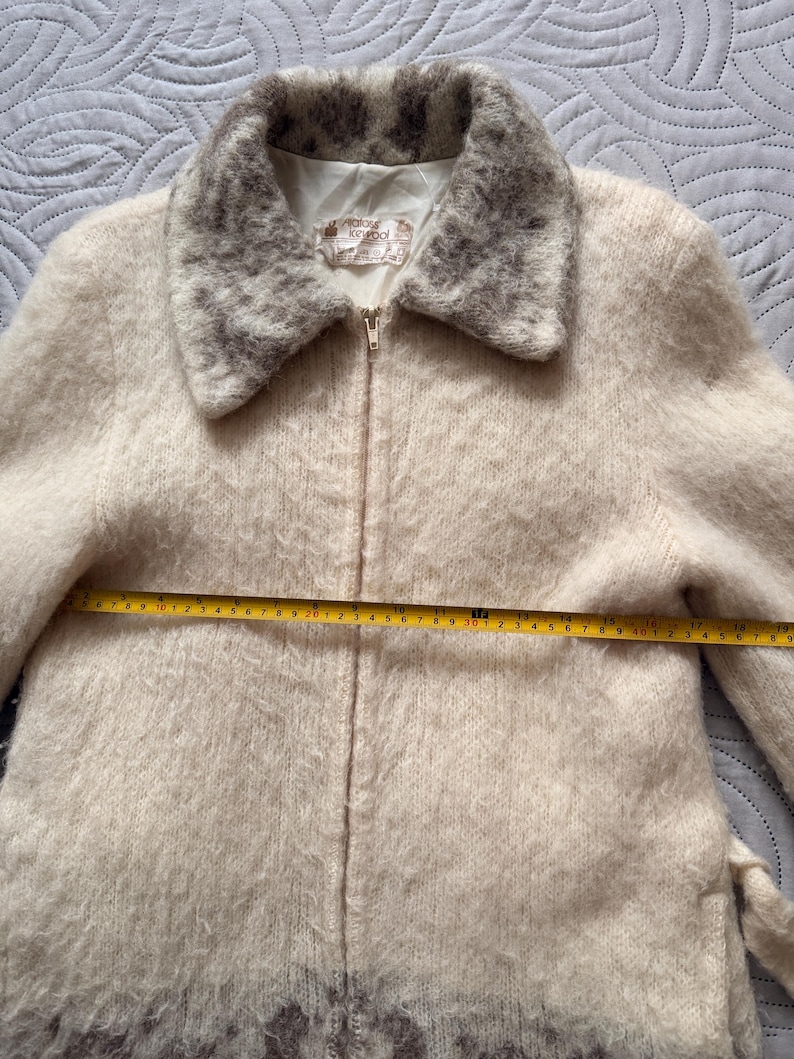 May include: Cream-colored vintage jacket with a fuzzy texture and a zip-up front. The collar and lower hem feature a patterned design in shades of gray. The jacket has a measurement of approximately 19 inches across the chest.