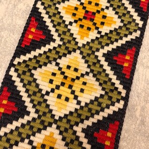 Puede incluir: Primer plano de un textil tejido con un patrón geométrico. El diseño presenta formas de diamante repetidas en amarillo, rojo, negro, blanco y verde oliva. El textil tiene un borde negro y una apariencia texturizada.