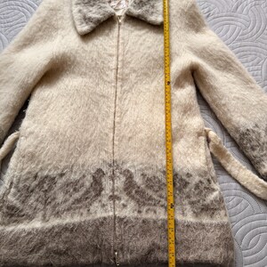 May include: A cream-colored, zip-up coat with a fuzzy texture and a wide collar. The coat features a brown patterned design at the bottom and a matching belt. The coat measures approximately 30 inches in length.