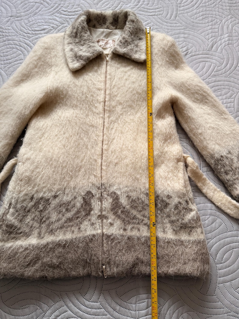 May include: A cream-colored, zip-up coat with a fuzzy texture and a wide collar. The coat features a brown patterned design at the bottom and a matching belt. The coat measures approximately 30 inches in length.