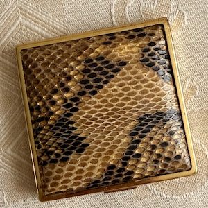 Może przedstawiać: Kwadratowy kompakt z ramką z metalu w kolorze złotym. Wierzch kompaktu zdobi wzór skóry węża w odcieniach brązu, beżu i czerni. Kompakt jest zamknięty i leży na kremowej tkaninie.