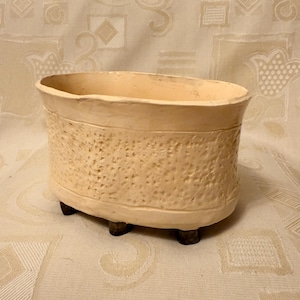 Könnte beinhalten: Cremefarbener, oval geformter Keramik-Blumentopf mit strukturierter Außenseite und drei kleinen, dunkelbraunen Füßen. Der Topf hat einen glatten Rand und ist für den Innen- oder Außenbereich konzipiert. Die neutrale Farbe ergänzt verschiedene Einrichtungsstile.