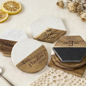 May include: A collection of personalized coasters with wood and white marble accents. The coasters feature engraved names and dates, with text such as "The Jordan Est. 2026" and "The Martinez Est. 2026". The coasters are in various geometric shapes.