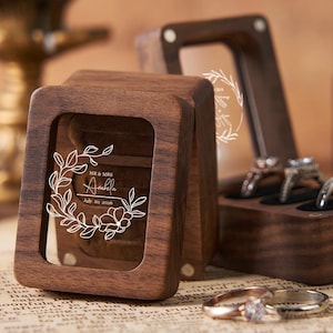 May include: Wooden ring boxes with clear windows, one open and one closed, displaying rings. The open box holds three rings, while the closed box has a floral design and the names "Mr & Mrs Anelle" and the date "July 20, 2026".