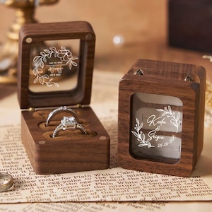 Può includere: Due scatole portanelli in legno con finestre in acrilico trasparente. Una scatola è aperta, mostrando un anello di diamanti. L'altra scatola è chiusa. Entrambe le scatole hanno disegni floreali incisi e nomi. Su sfondo di carta vintage.