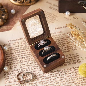 May include: A wooden ring box with a clear lid, open to reveal three rings with sparkling stones. The box sits on a vintage paper background with pearls and dried flowers. The box is approximately 4 inches long.