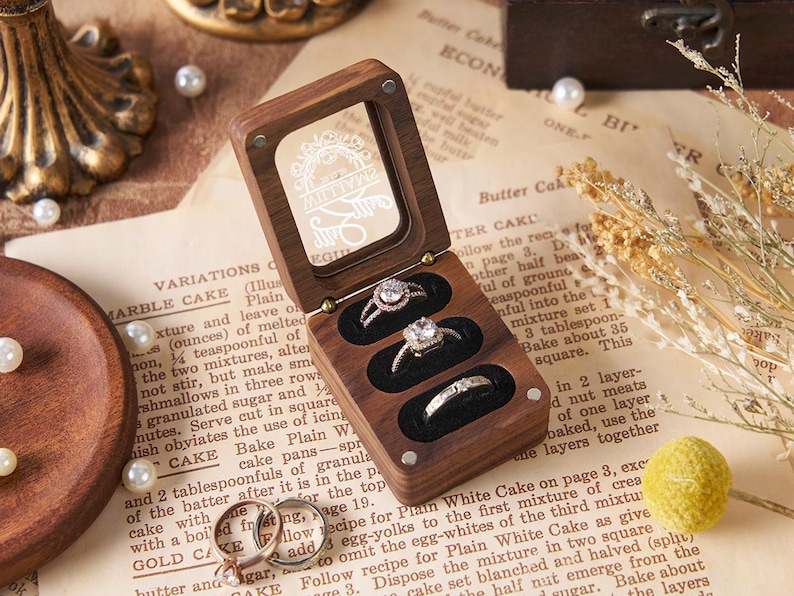 May include: A wooden ring box with a clear lid, open to reveal three rings with sparkling stones. The box sits on a vintage paper background with pearls and dried flowers. The box is approximately 4 inches long.