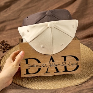 May include: A wooden storage box featuring the black text DAD with the names Jackson, Adelaa, and Crystal separated by heart icons. Two baseball caps, one dark grey and one off-white, are resting inside the box, which has a light wood grain finish.