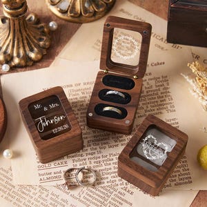 May include: Wooden ring boxes with clear lids, some open to display rings. One box is engraved with "Mr. & Mrs. Johnson 12.26.26". The boxes sit on vintage paper with a floral arrangement and pearls.