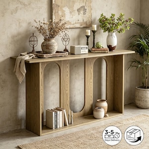 Può includere: Un tavolo console in rovere chiaro caratterizzato da due ampie aperture ad arco nella base. Il piano superiore ospita un vaso in ceramica con fiori secchi, due sculture a sfera in metallo, una radio, due candele e un vaso bianco con fogliame.