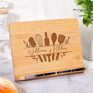May include: A rectangular bamboo cookbook stand featuring a laser-engraved design of kitchen utensils including a spatula, whisk, rolling pin, spoon, and turner, with the text "William's Kitchen" centred and two black metal page holders.