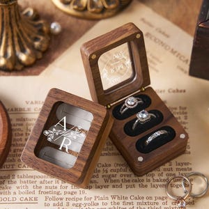 May include: Wooden ring boxes with clear lids, showcasing engagement rings. One box is open, displaying three rings with sparkling stones. The other box is closed, with the initials "AR" and floral design. The boxes sit on a vintage paper background.