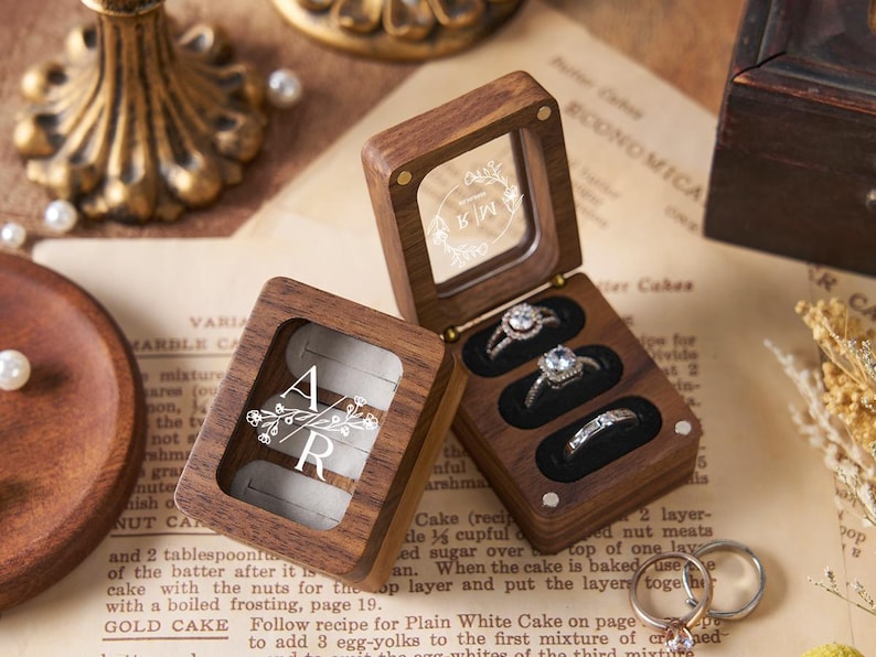 May include: Wooden ring boxes with clear lids, showcasing engagement rings. One box is open, displaying three rings with sparkling stones. The other box is closed, with the initials "AR" and floral design. The boxes sit on a vintage paper background.