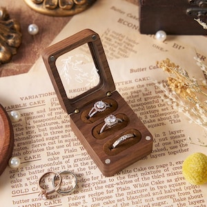 May include: A wooden ring box with a clear lid, holding three diamond rings. The box sits on a vintage recipe page, surrounded by pearls and decorative elements. The rings are set in brown velvet.