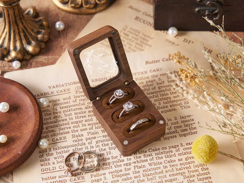 May include: A wooden ring box with a clear lid, holding three diamond rings. The box sits on a vintage recipe page, surrounded by pearls and decorative elements. The rings are set in brown velvet.