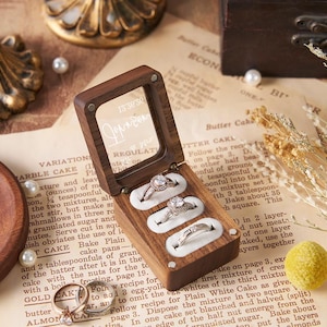 May include: A wooden ring box with a clear lid, holding three diamond rings. The box sits on vintage paper with scattered pearls and a wooden plate. A gold-colored decorative stand is in the background.