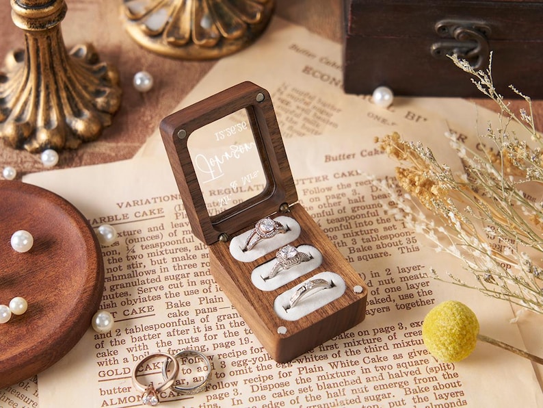 May include: A wooden ring box with a clear lid, holding three diamond rings. The box sits on vintage paper with scattered pearls and a wooden plate. A gold-colored decorative stand is in the background.