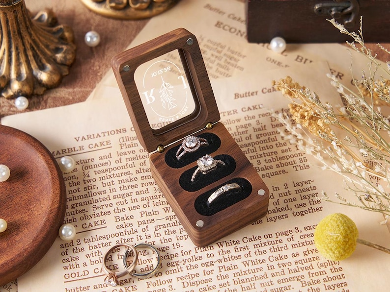 May include: A wooden ring box with a clear lid, holding three rings with sparkling stones. The box is open, revealing the rings against a black velvet interior. The box is on a vintage paper background.