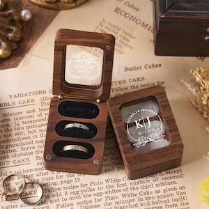 May include: Wooden ring boxes with clear lids, one open with three rings displayed. The other box has a personalized design with initials and a date. The boxes sit on vintage paper.