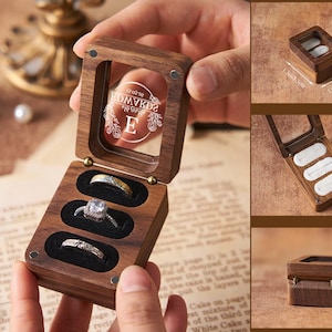 May include: A wooden ring box with a clear lid, holding three rings. The box is open, revealing the rings in black velvet slots. The lid has etched text and a decorative design. The box is 1.9 inches long, 1.9 inches wide, and 0.9 inches high.