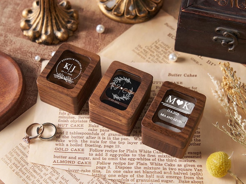 May include: Three wooden ring boxes with clear tops, each engraved with initials and dates. The boxes are on a vintage paper background, with wedding rings visible. Decorative elements include pearls and a gold-colored stand.
