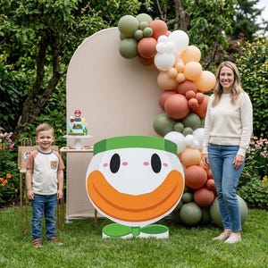 Puede incluir: Un recorte de cartón grande y plano de un personaje tipo payaso de color verde, blanco y naranja con una amplia sonrisa, colocado frente a un telón de fondo arqueado de color beige decorado con globos verde salvia, terracota y blancos.