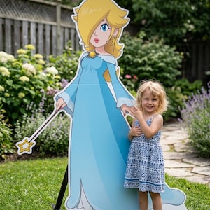 Puede incluir: Un gran recorte de cartón azul claro de una princesa de dibujos animados con corona y varita, sobre un césped verde. Una niña pequeña con un vestido azul estampado está de pie junto al recorte, sonriendo.