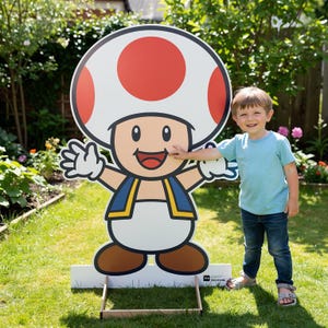 May include: A large, colorful cardboard cutout of the character Toad from the Super Mario video game series. Toad has a white mushroom cap with red spots, a blue vest, and brown shoes. A young child is standing next to the cutout.