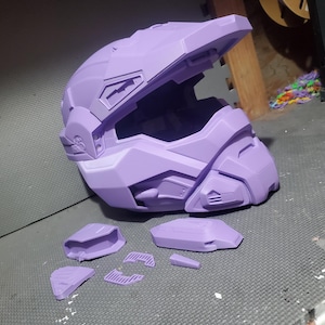 May include: A lavender-colored, full-face tactical helmet with a prominent visor and angular, geometric plating. Several smaller, detached lavender plastic components are arranged in front of the helmet, showing a modular design.