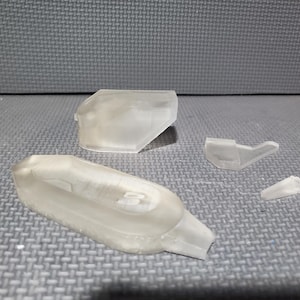 Könnte beinhalten: Mehrere durchscheinende Kunststoffkomponenten liegen auf einer grauen Oberfläche. Die Teile scheinen Teile eines größeren Objekts zu sein, möglicherweise ein Modell oder ein Prototyp. Die Formen variieren.