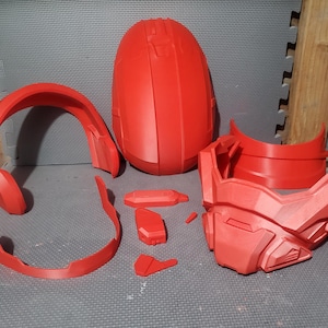 May include: A disassembled red helmet, likely for cosplay or a costume. The components include a helmet shell, faceplate, neck guard, and smaller pieces. The parts are arranged on a gray surface.