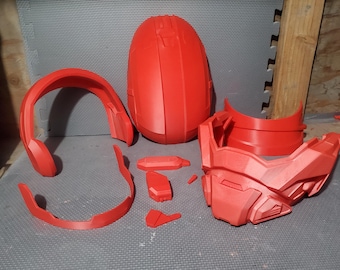 May include: A collection of red helmet components, possibly for a costume or cosplay. The parts include a helmet shell, faceplate, neck guard, and smaller pieces. They are arranged on a grey surface.