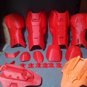 Op de afbeelding: Een verzameling rode en oranje 3D-geprinte pantseronderdelen, waaronder been- en armbeschermers, borstplaten en kleinere componenten. De stukken zijn gerangschikt op een blauw oppervlak en tonen de verschillende onderdelen van een kostuum of rekwisiet.
