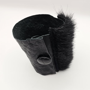 Pode incluir: Uma pulseira de punho de couro preto com uma superfície texturizada e acabamento em pele preta. A pulseira tem um fecho de botão preto. Um acessório elegante.