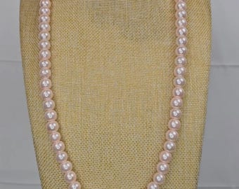 May include: A light pink pearl necklace on a mannequin. The necklace has graduated sizes of round pearls, with the largest pearls in the centre. The pearls have a soft, lustrous sheen, and the necklace is displayed against a neutral background.
