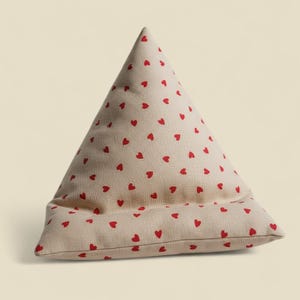 Peut inclure: Un coussin triangulaire beige orné de petits cœurs rouges. Le coussin est doté d'une poche pour maintenir un livre ou une tablette. Le tissu a un aspect texturé, et les cœurs sont répartis uniformément sur la surface.