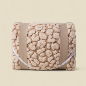 Peut inclure: Une couverture beige roulée avec un motif léopard texturé. La couverture est enveloppée d'une bande de papier brun et attachée avec un ruban de dentelle blanc. La couverture est de couleur neutre et convient à une chambre ou un salon.