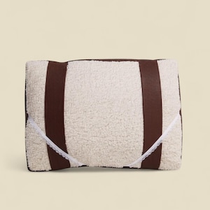 Peut inclure: Un coussin rectangulaire avec une surface texturée de couleur crème. Deux lanières verticales en cuir marron foncé descendent sur le devant. Une bordure en dentelle blanche est cousue sur les côtés. Accessoire de décoration pour la maison.
