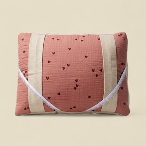 Può includere: Un cuscino rettangolare color rosa cipria con un motivo a cuore. Il cuscino ha due strisce verticali di tessuto color crema e un bordo in pizzo bianco. Il cuscino è progettato per uso decorativo o funzionale.